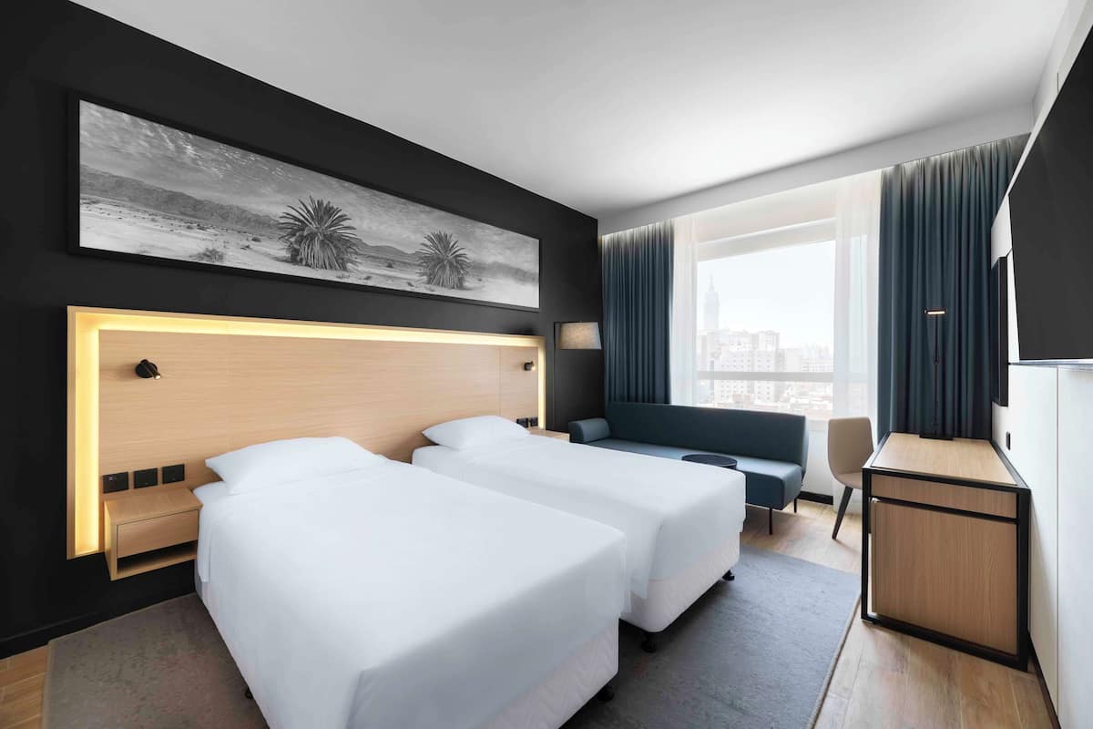 A hotel room with two beds, a desk, a sofa, a large window with curtains, and a city view.