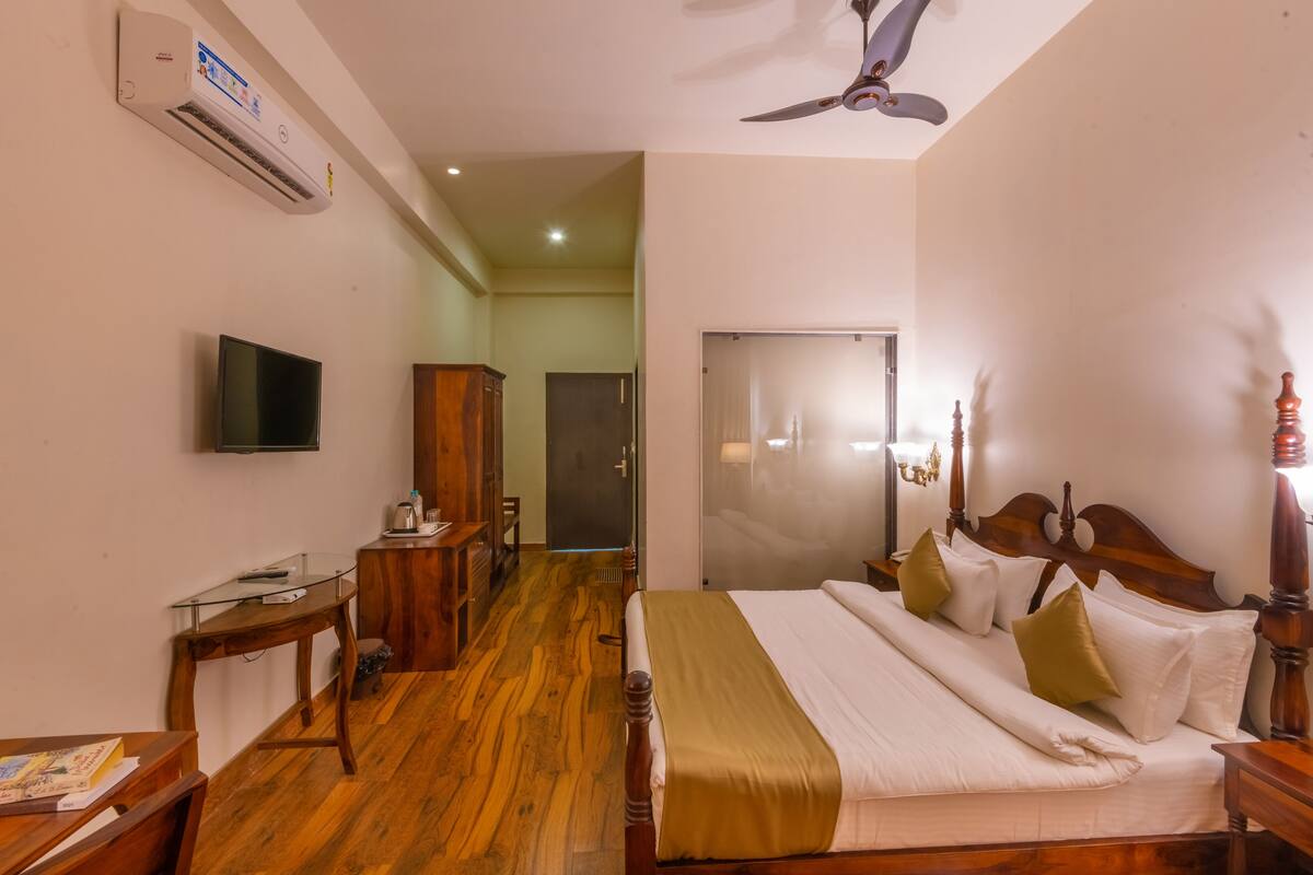 A hotel room with a wooden floor, a bed with white and gold bedding, a television on the wall, and a ceiling fan.