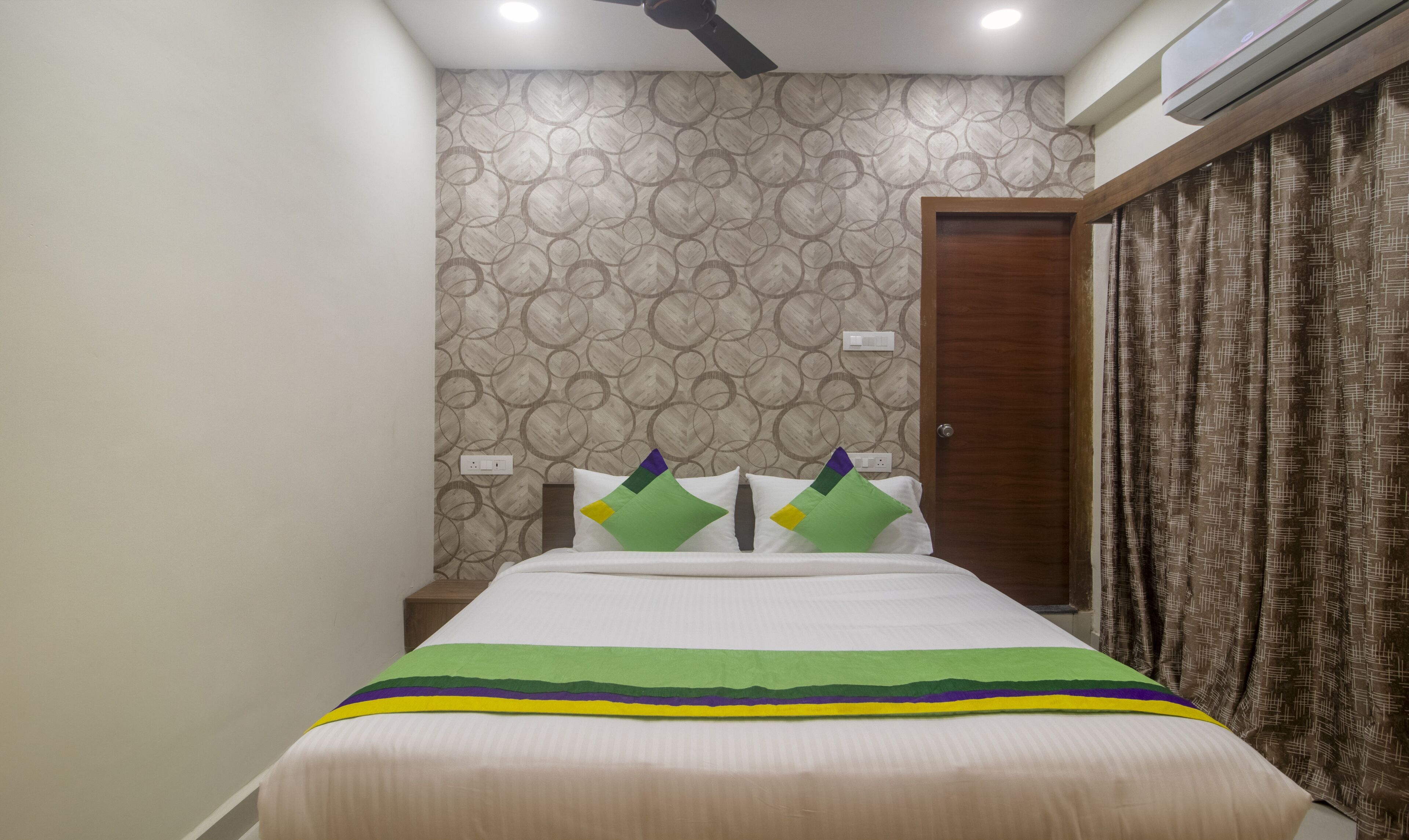 A bedroom with a bed, two pillows, a nightstand, a wall with a circular pattern, a door, and an air conditioning unit.