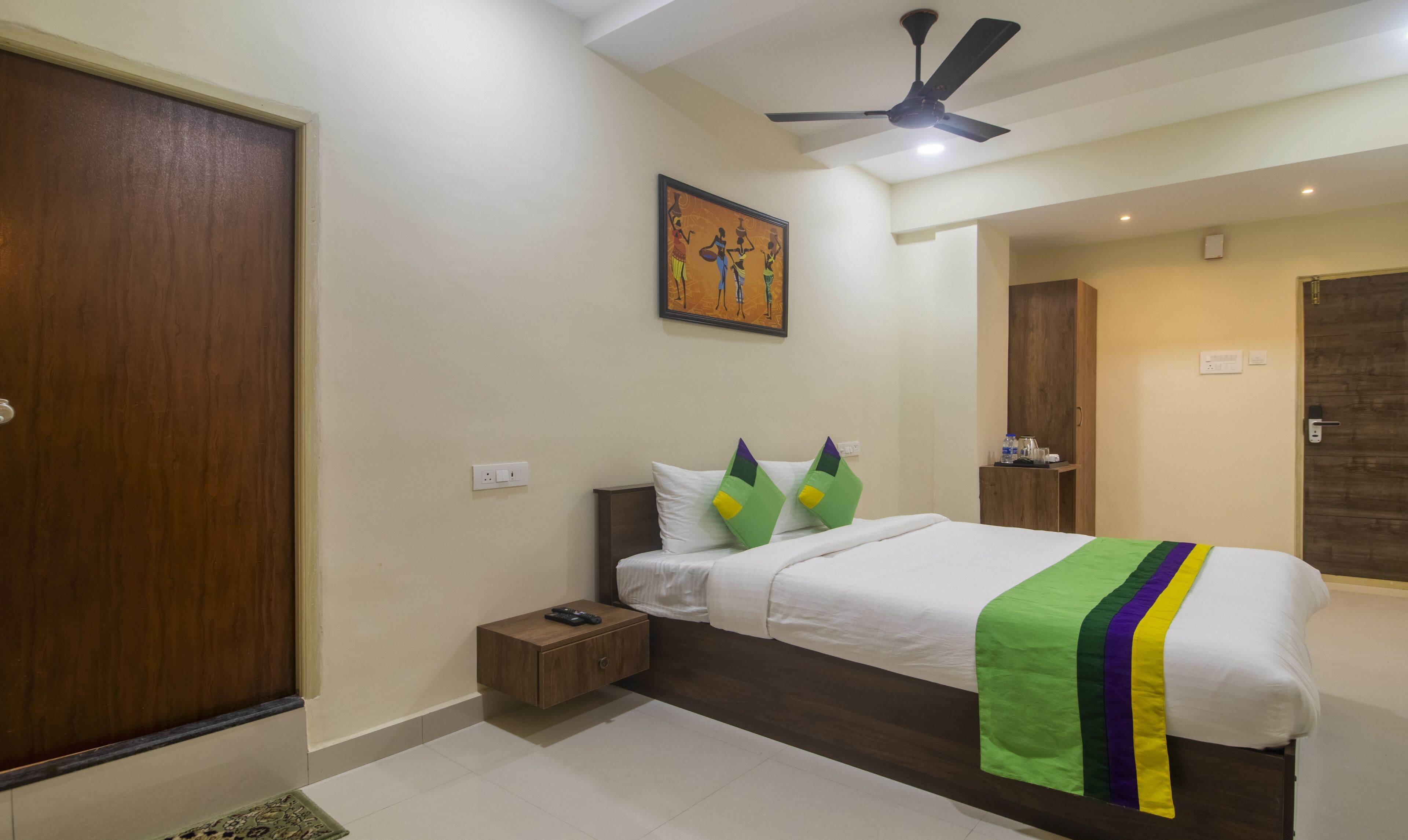 A hotel room with a bed, a nightstand, a ceiling fan, a painting on the wall, and a wardrobe.