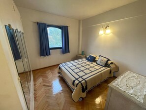 Un dormitorio con cama, mesita de noche, ventana con cortinas y un aplique de pared.
