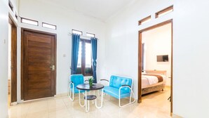A small, well-lit room with a wooden door, a blue sofa, a small round table with a plant, and a bed with a TV in the background. - FHStay Maguwo (Depok)