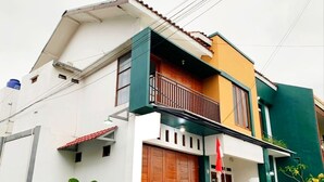 A two-story house with a balcony, a wooden door, and a small garden area with plants. - FHStay Maguwo (Depok)