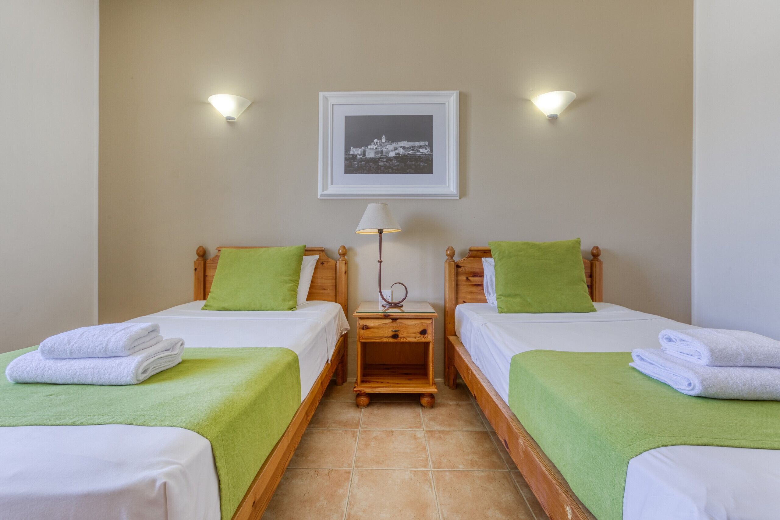 A hotel room with two single beds, each with a green pillow and a white towel. There is a nightstand with a lamp between the beds, and a framed picture on the wall above.