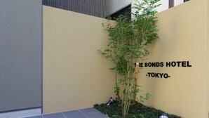 Entrada de un hotel con un letrero que dice "The Bonds Hotel - Tokyo" y un pequeño jardín con un árbol y arbustos.