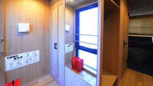 Habitación de hotel con paredes de madera, una ventana con persianas y una maleta roja sobre un estante, con un letrero que indica un dispositivo ORIRO.