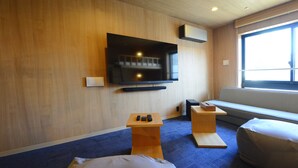Una habitación de hotel moderna con un televisor de pantalla plana grande montado en la pared, una pequeña mesa de madera con dos mandos a distancia, un banco y una cama con colchón blanco.