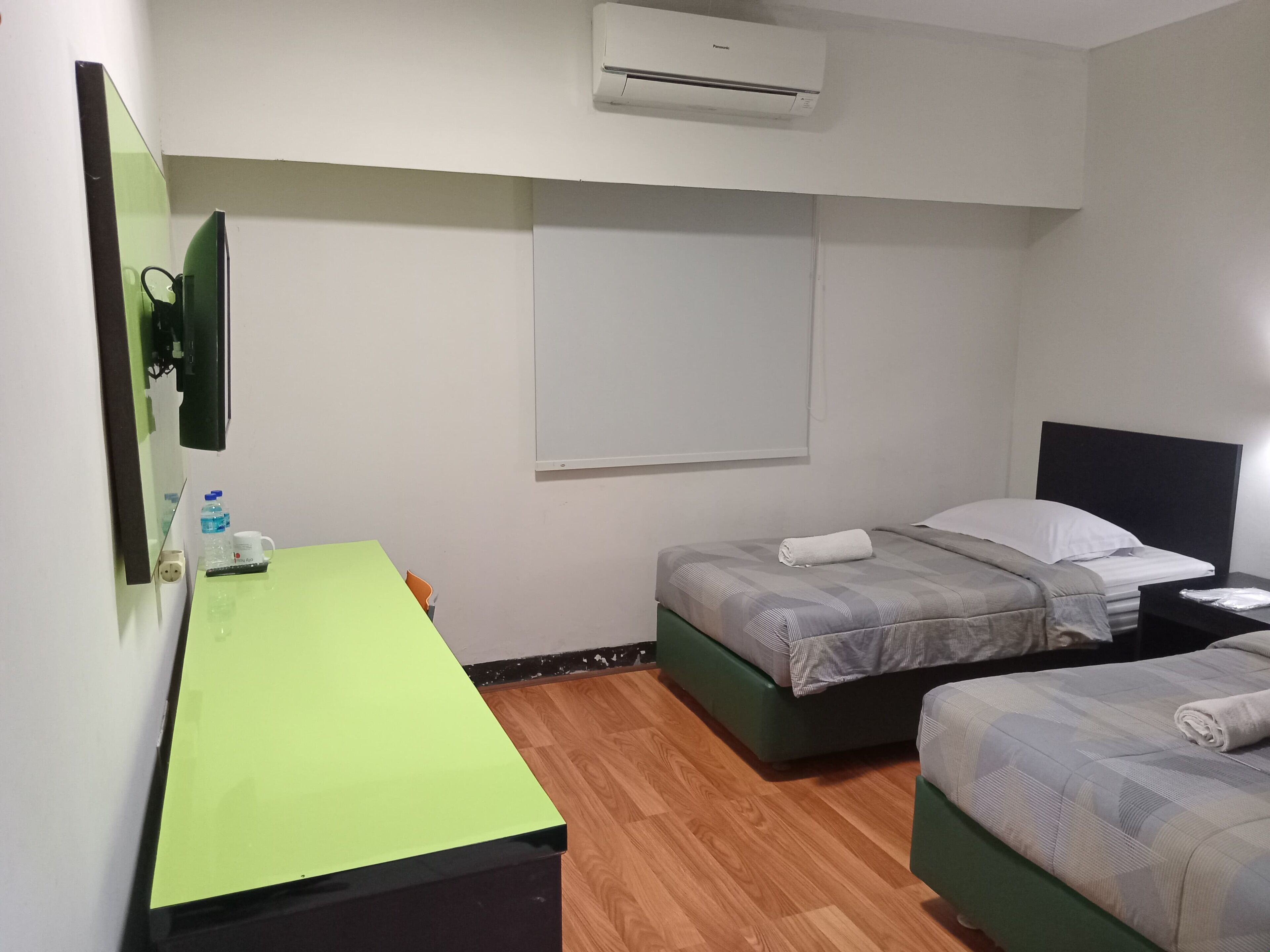 Una habitación de hotel con un escritorio verde, dos camas individuales, un aire acondicionado de pared y un espejo.