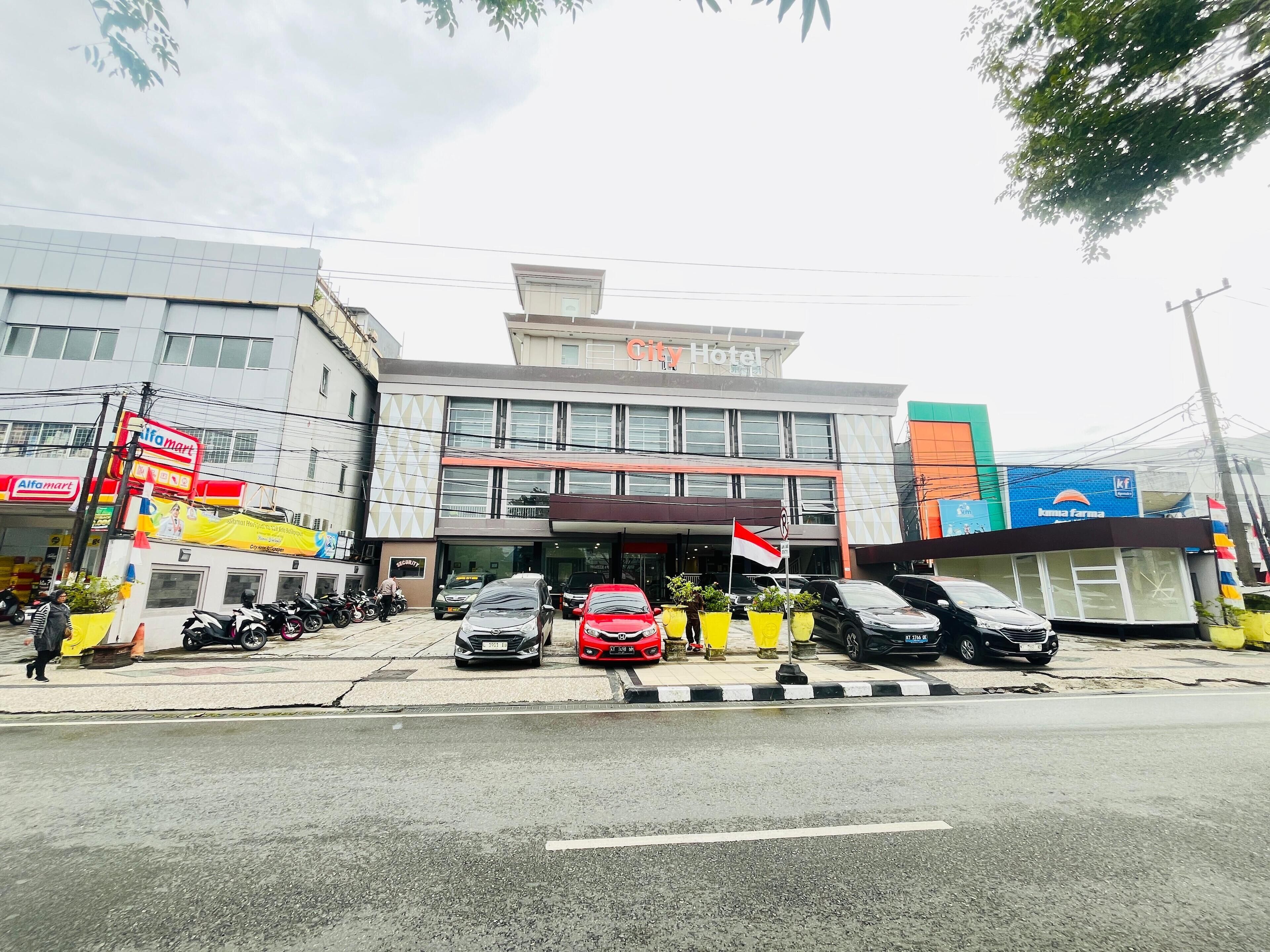 Un hotel con fachada moderna, entrada visible y un estacionamiento con varios coches y motocicletas.