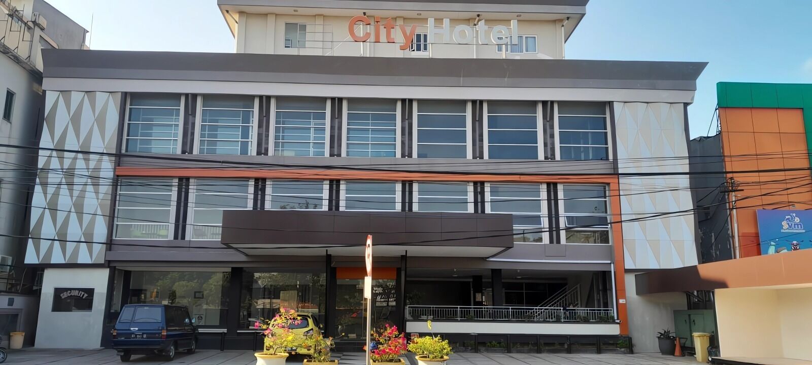 A modern hotel with a prominent 'City Hotel' sign, large windows, and a covered entrance area with potted plants.