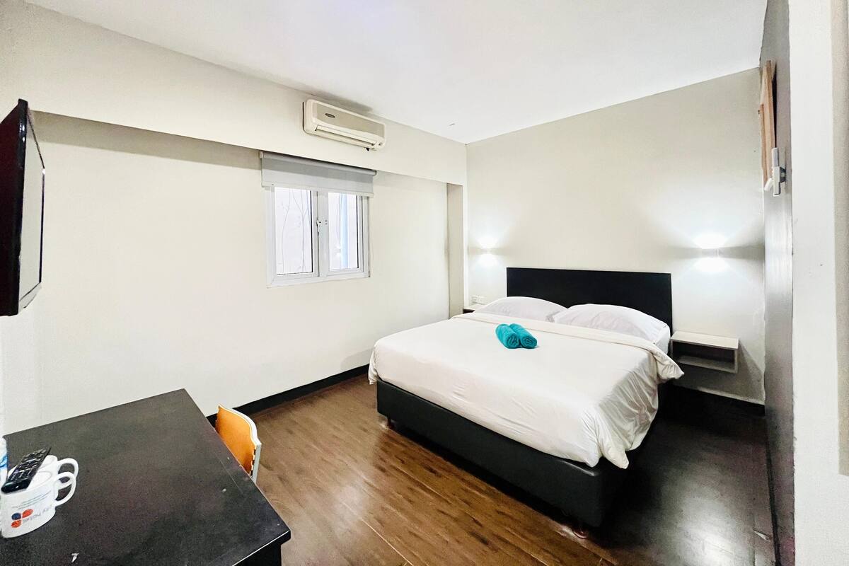 A hotel room with a bed, bedside lamps, a desk with a phone, and a window with blinds.