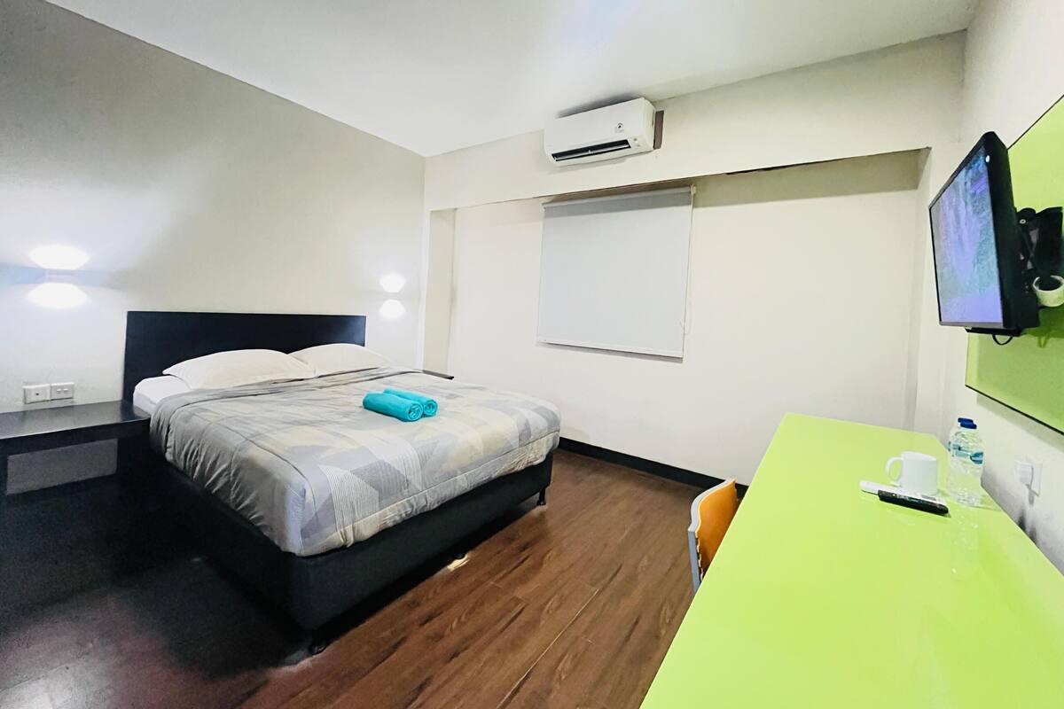 A hotel room with a bed, a desk, a chair, an air conditioner, a television, and a window with a blind.