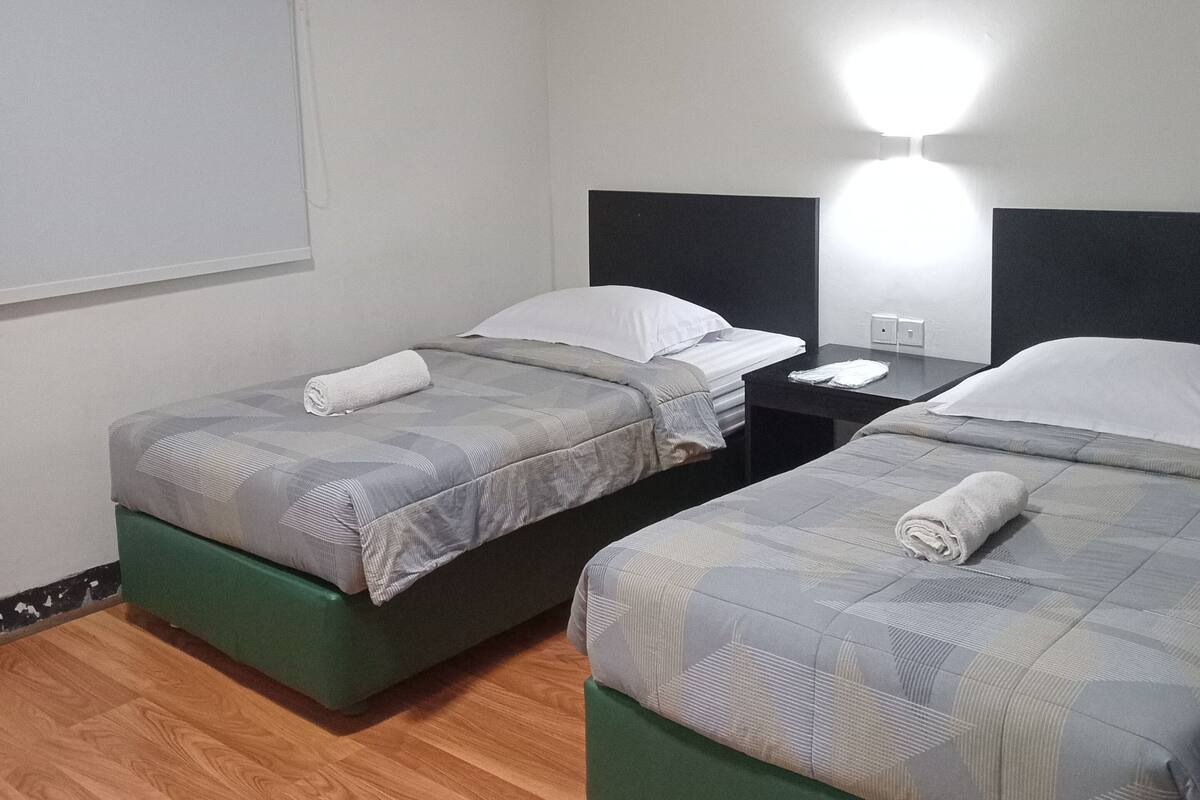 A hotel room with two beds, each with a pillow and a rolled towel. There is a nightstand with a lamp between the beds.