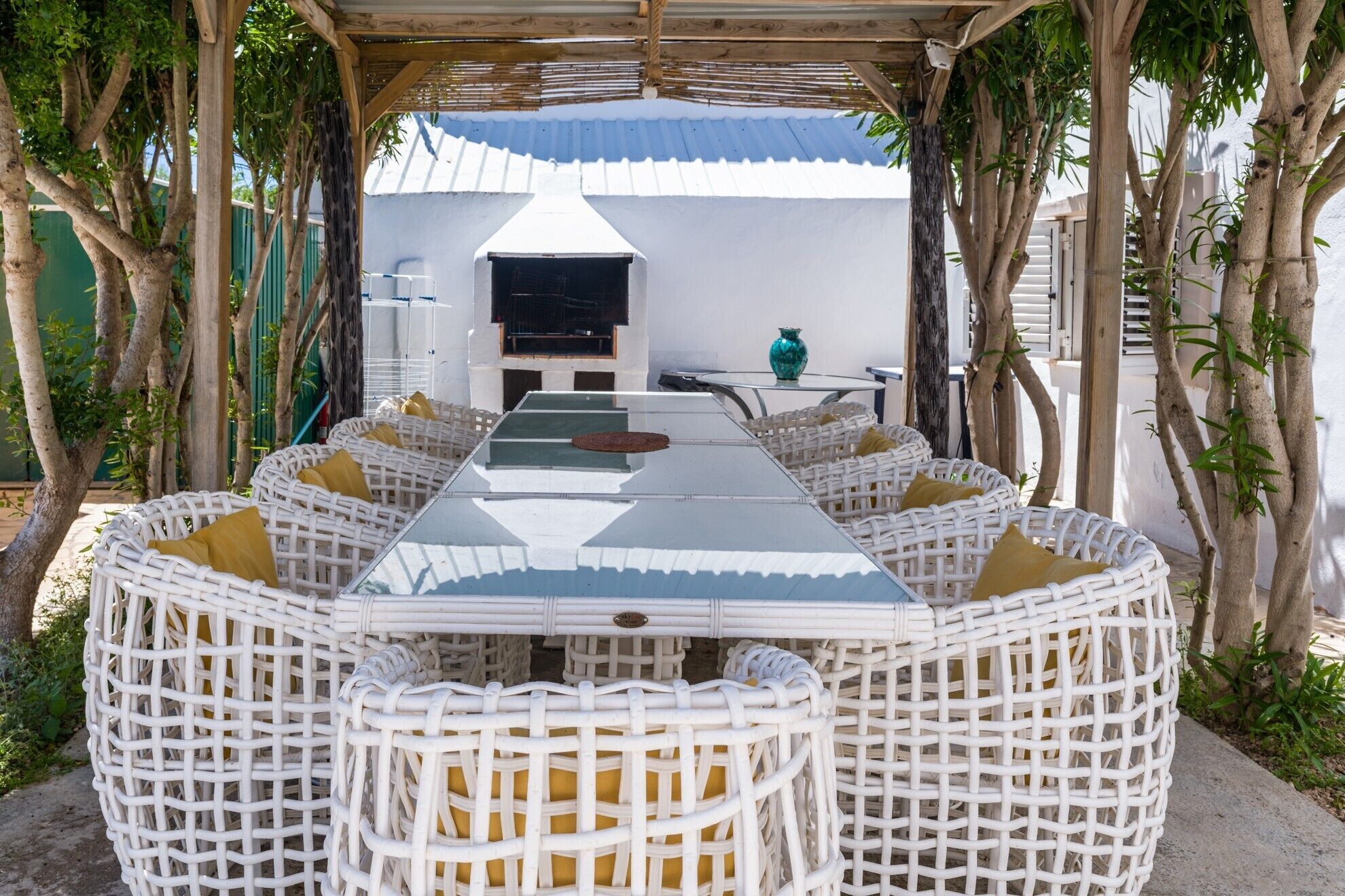 Un espacio de comedor al aire libre con techo, con una mesa larga y sillas de mimbre, rodeado de árboles.
