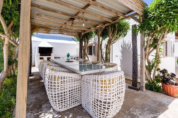 An outdoor seating area with a glass-top table and wicker chairs, surrounded by trees and a covered pergola.