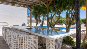 Un'area pranzo all'aperto riparata, con tavolo in vetro e sedie in vimini, circondata da alberi e una piscina.