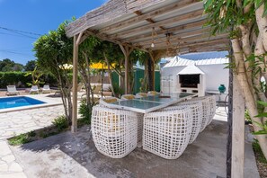A covered outdoor dining area with a pool, white wicker furniture, and a grill.
