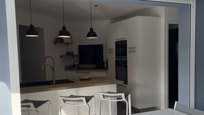 Eine moderne Küche mit weißen Schränken, einer zentralen Kochinsel und Pendelleuchten. Es sind drei weiße Barhocker sowie ein Esstisch mit Stühlen vorhanden.