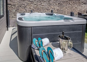 A hot tub with a stone wall backdrop, a bottle of champagne, and flip-flops on a tiled deck.