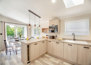 A modern kitchen with wooden cabinets, a built-in microwave, a large window with blinds, and a dining area with chairs and a table.