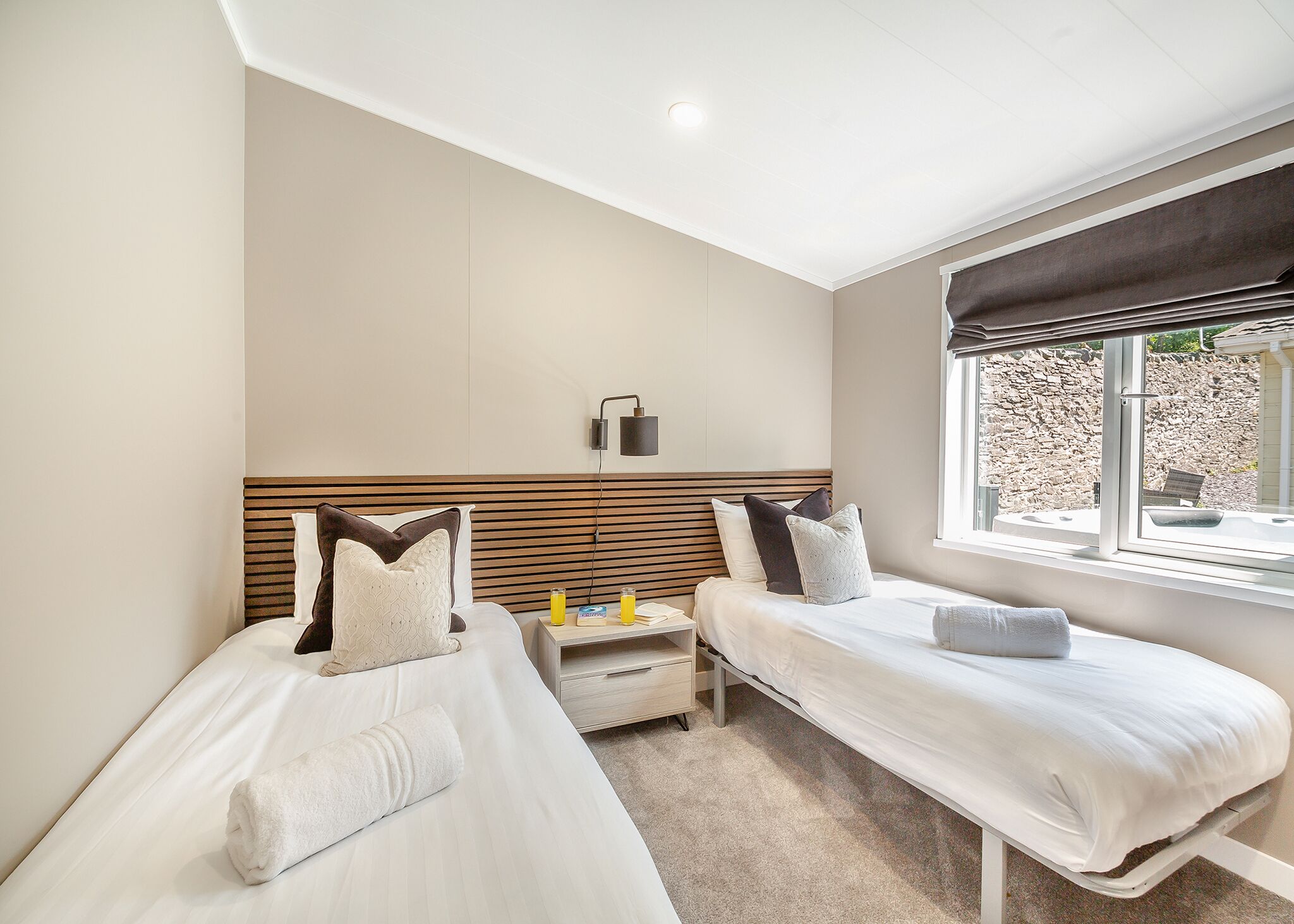 A hotel room with two beds, a bedside table with a lamp, a window with a view of a stone wall and a hot tub, and a ceiling light.