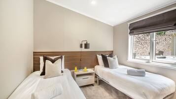A hotel room with two beds, a bedside table with a lamp, a window with a view of a stone wall and a hot tub, and a ceiling light.