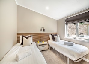 A hotel room with two beds, a bedside table with a lamp, a window with a view of a stone wall and a hot tub, and a ceiling light.