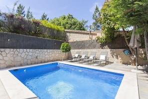 A rectangular swimming pool with a stone wall and a small tree in the corner.