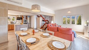 Una cocina moderna con una isla central, un comedor con mesa de madera dispuesta para cuatro, y una escalera que lleva a un dormitorio tipo loft.