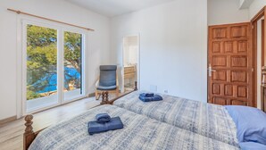 Un dormitorio con dos camas, una silla, una puerta de madera y vistas a una piscina a través de amplios ventanales.