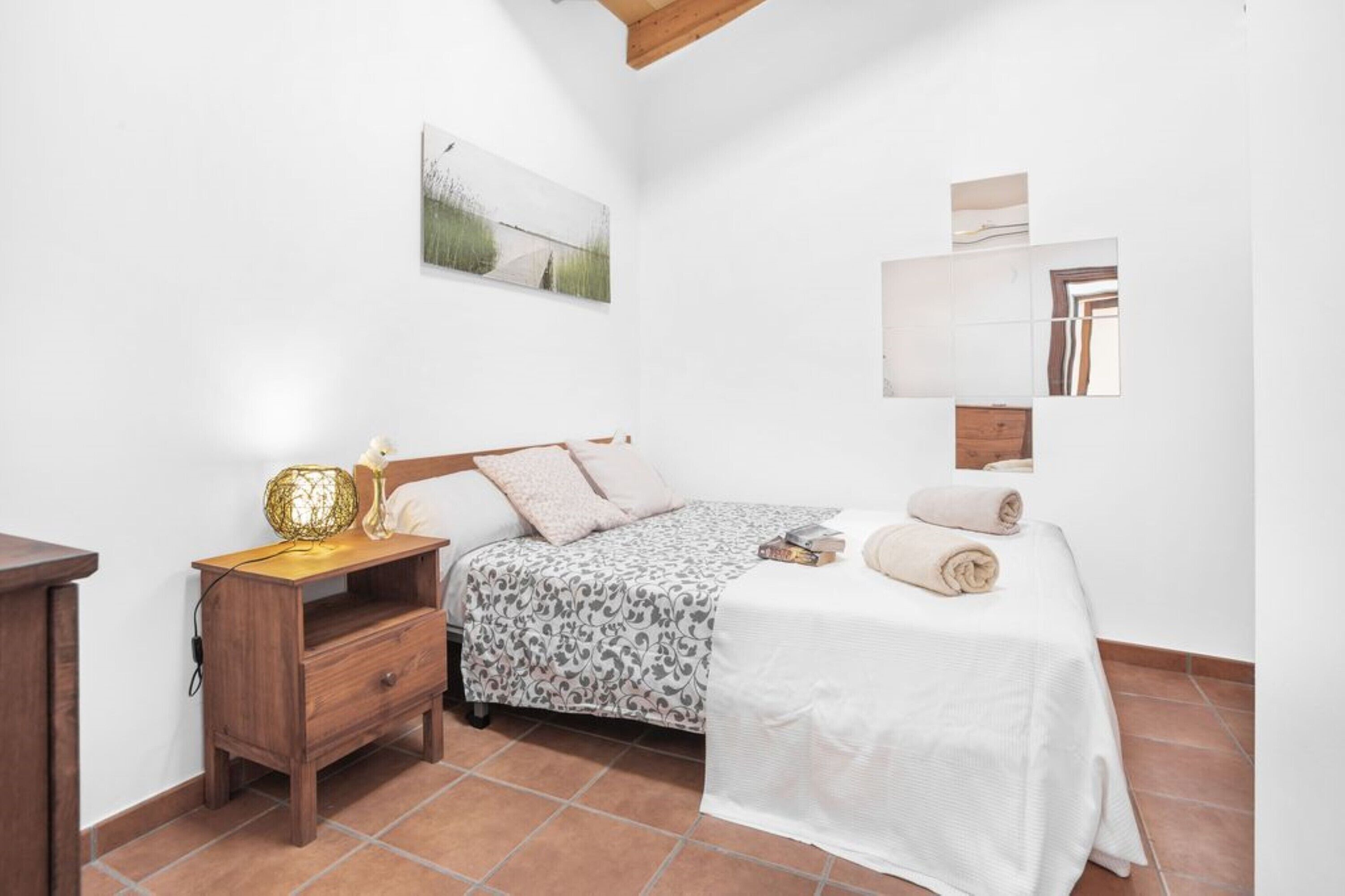 A bedroom with a bed, a nightstand, a lamp, a mirror, and a painting on the wall.