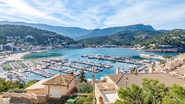 Un puerto costero con barcos, edificios y montañas al fondo.
