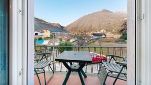 A balcony with a table and chairs, a red car, and a view of a town and mountains.