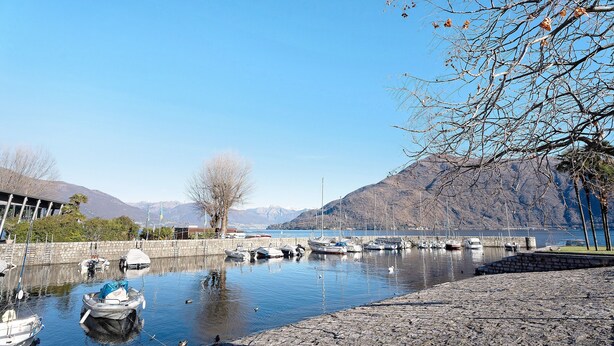 A marina with boats docked, a stone walkway, and mountains in the background.