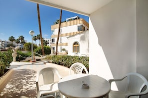 A balcony with white chairs and a table, overlooking a garden with palm trees and a building with balconies.