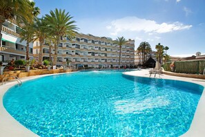 Ein Swimmingpool mit klarem, blauem Wasser, umgeben von Palmen und einem mehrstöckigen Hotelgebäude.