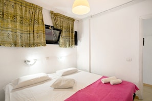 A hotel room with a bed, pillows, a pink blanket, a television mounted on the wall, and a window with curtains.