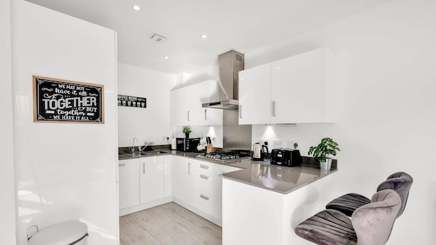A modern kitchen with white cabinets, a stainless steel range hood, and a bar area with a bar stool. - Foxherne 6 Bdrm, 3BA Hse -17 (Slough)