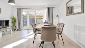 A modern living room with a glass dining table, comfortable seating, a flat-screen TV, and a large window with curtains. - Foxherne 6 Bdrm, 3BA Hse -17 (Slough)