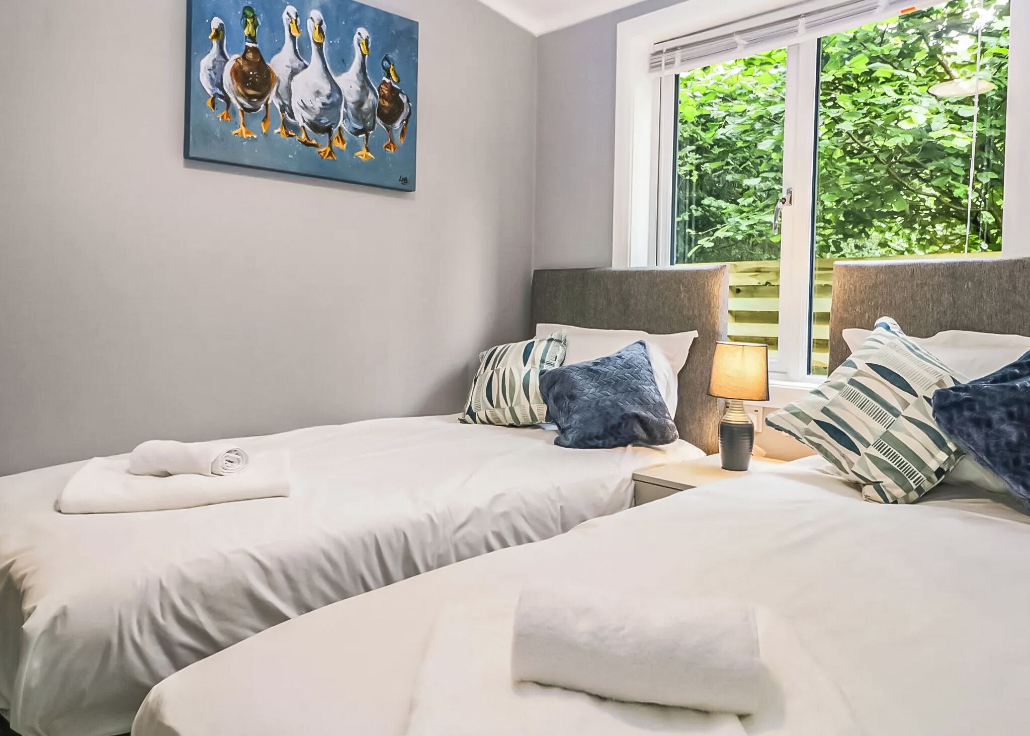 A hotel room with two beds, a painting of ducks, a window with a view of greenery, and a lamp on the nightstand.