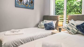 A hotel room with two beds, a painting of ducks, a window with a view of greenery, and a lamp on the nightstand.