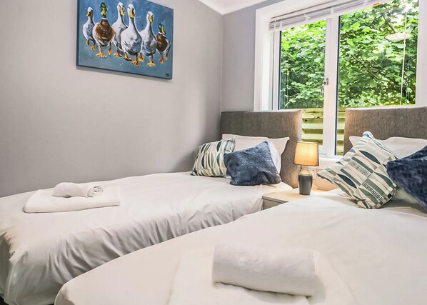 A hotel room with two beds, a painting of ducks, a window with a view of greenery, and a lamp on the nightstand.