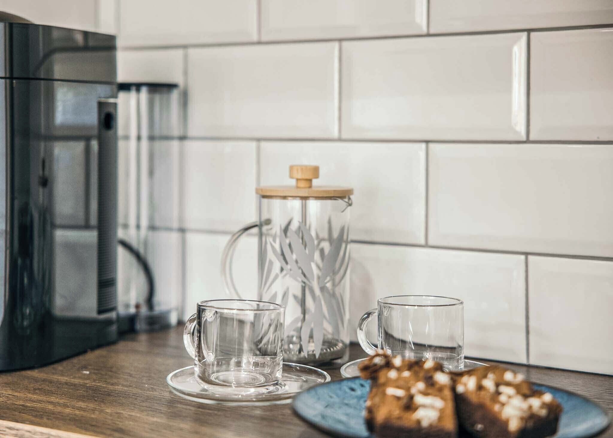 A kitchen counter with a coffee maker, two clear glasses, a glass jar with a wooden lid, and a plate with muffins.
