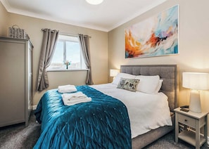 A hotel room with a bed, a nightstand, a lamp, a window with curtains, and a painting on the wall.