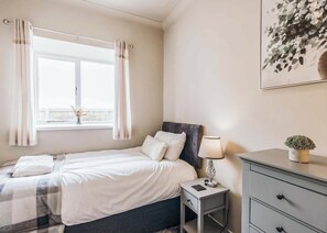Un dormitorio con cama, mesita de noche con lámpara, cómoda y ventana con cortinas.