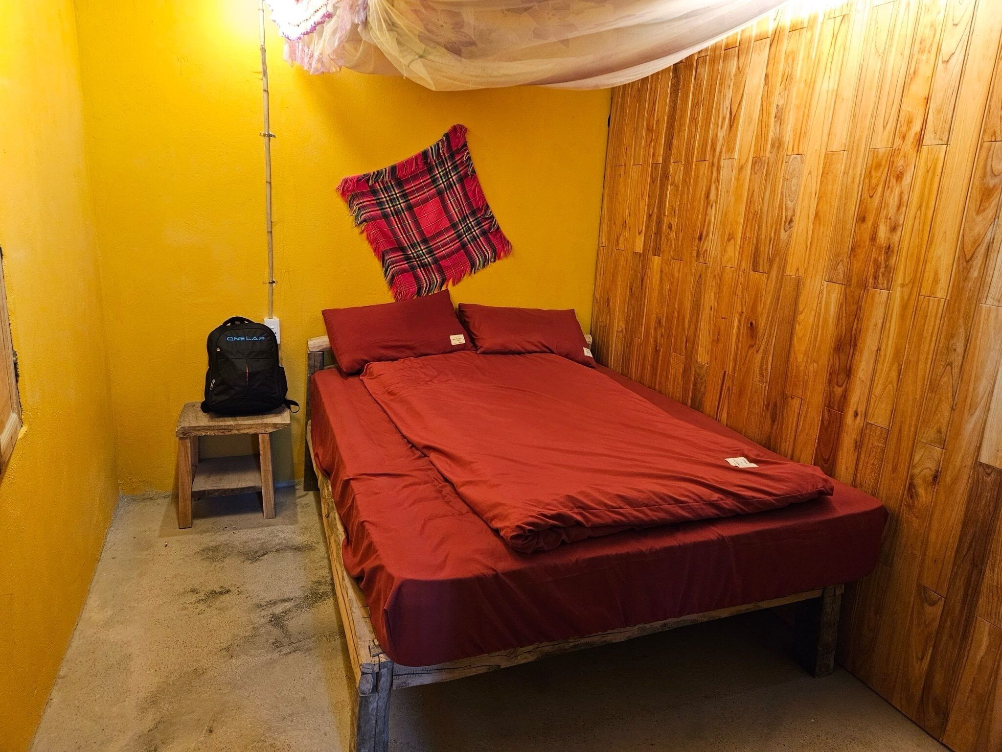 Une petite pièce comprenant un lit, une armoire en bois, un petit tabouret et un sac à dos.