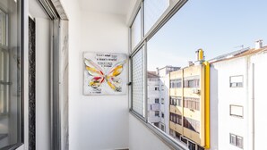 Un balcón estrecho con vista a edificios cercanos, una obra de arte con forma de mariposa montada en la pared y una ventana con una tubería amarilla.