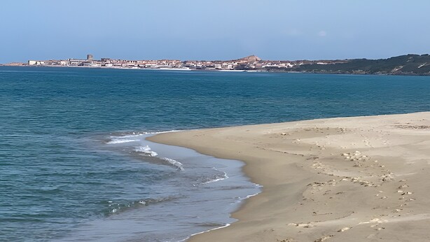 A beach with clear water, a sandy shore, and a distant town with buildings. - Holiday Home "Casa OriMar 419" with Shared Pool and Wi-Fi (Badesi)