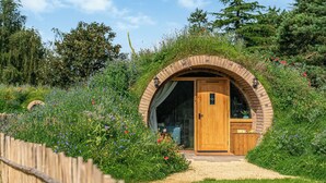 Une structure ronde en bois, dotée d’une porte, entourée d’un jardin où poussent diverses plantes et fleurs.