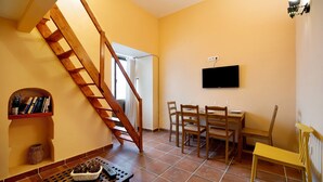Ein Zimmer mit einer Holztreppe, einem Esstisch mit Stühlen, einem an der Wand montierten Fernseher und einem Regal mit Büchern und Dekoration.